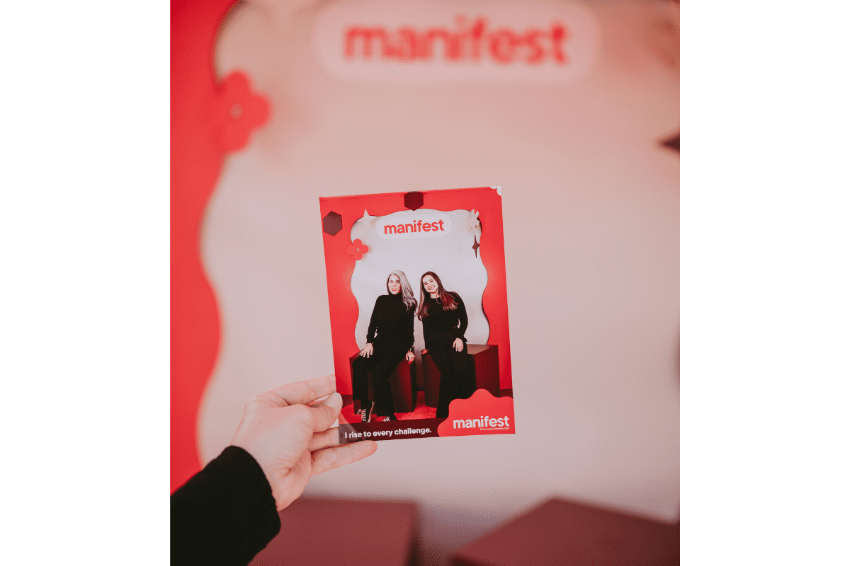 Hand holding a printed photo from a corporate photo booth activation at the Manifest event in Phoenix, Arizona. The branded red photo backdrop and printout feature two women seated in front of the Manifest logo with the caption “I rise to every challenge.” Ideal example of Phoenix Arizona corporate photo booth rentals by a local woman-owned business.
