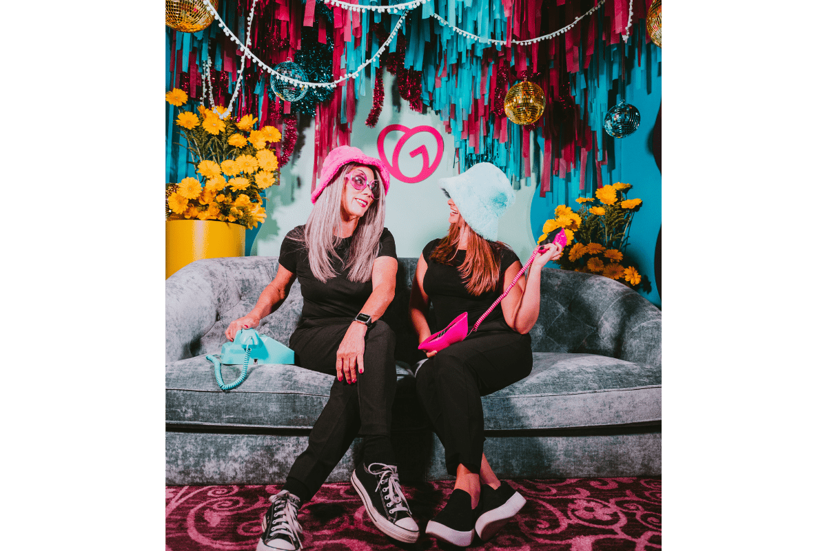 Group of female event photo booth attendants smiling and posing at a vibrant, woman-owned photo booth rental setup by MISGIF in Phoenix, Arizona featuring bold decor, a velvet couch, and colorful fringe backdrop—highlighting MISGIF’s creative, woman-led event rentals for corporate events.
